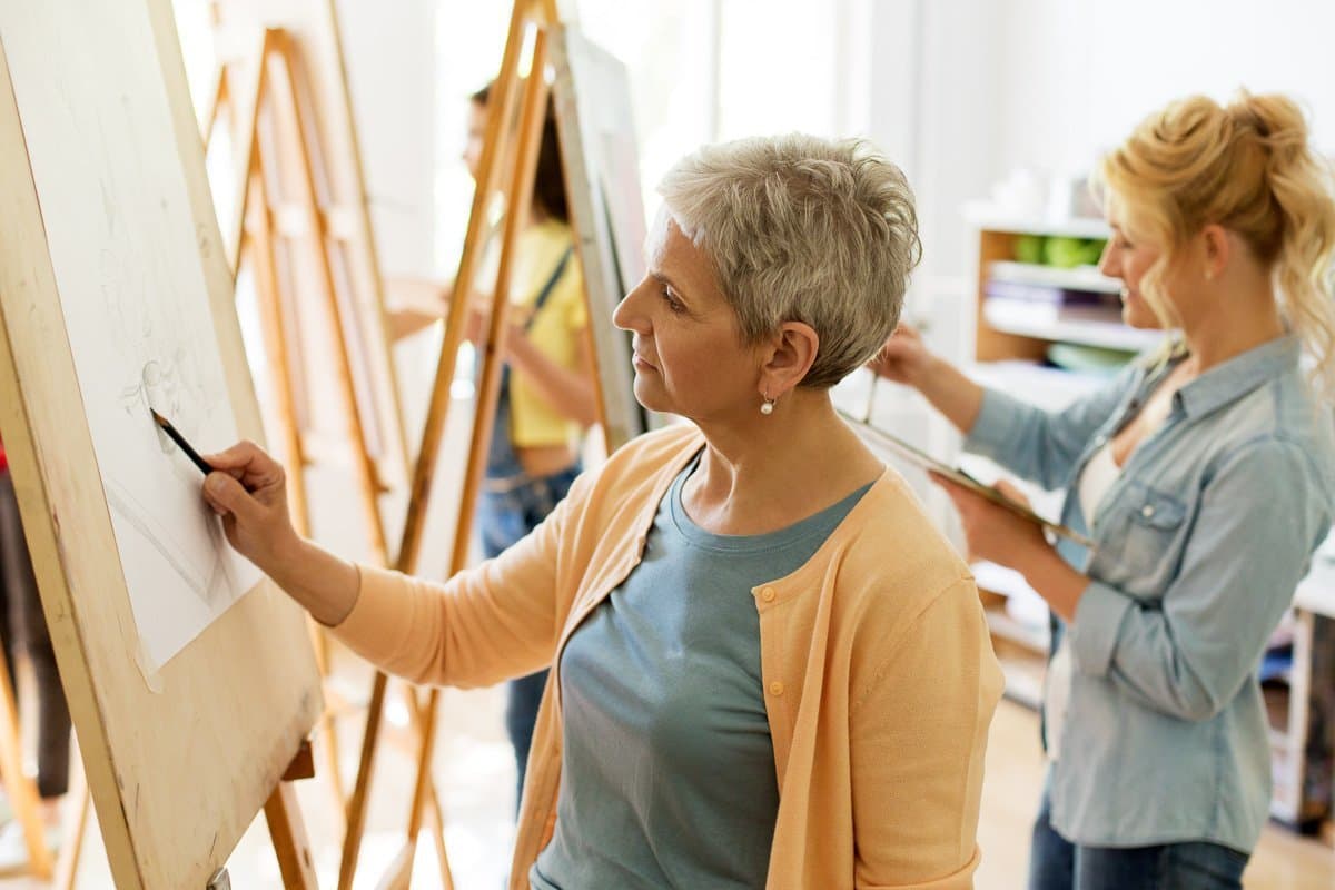 Two women drawing or sketching on easels in an art class or studio, with shelves and other students in the background. Varcity, Purdue University, Texas A&M University, retirement, on campus developments, campus life for life.