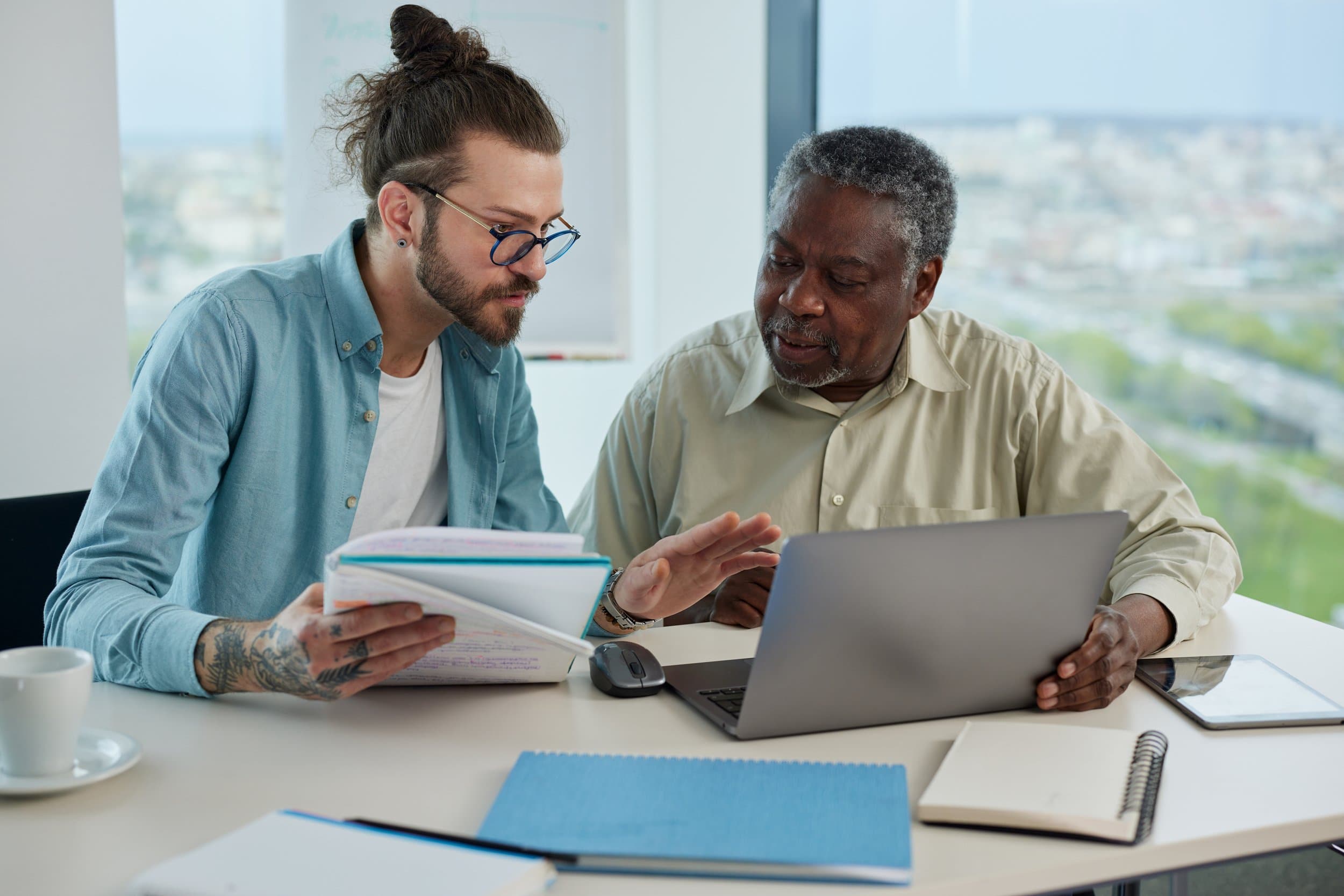 Two men are working together at a table with a laptop, notebooks, and a tablet, in an office with large windows showing a city view. Varcity, Purdue University, Texas A&M University, retirement, on campus developments, campus life for life.
