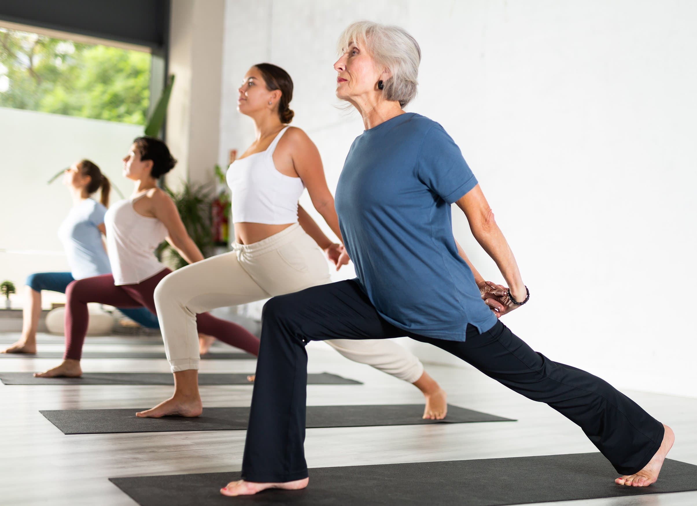Older woman and younger women practicing yoga in a studio, stretching in a lunge pose on black mats. Varcity, Purdue University, Texas A&M University, retirement, on campus developments, campus life for life.
