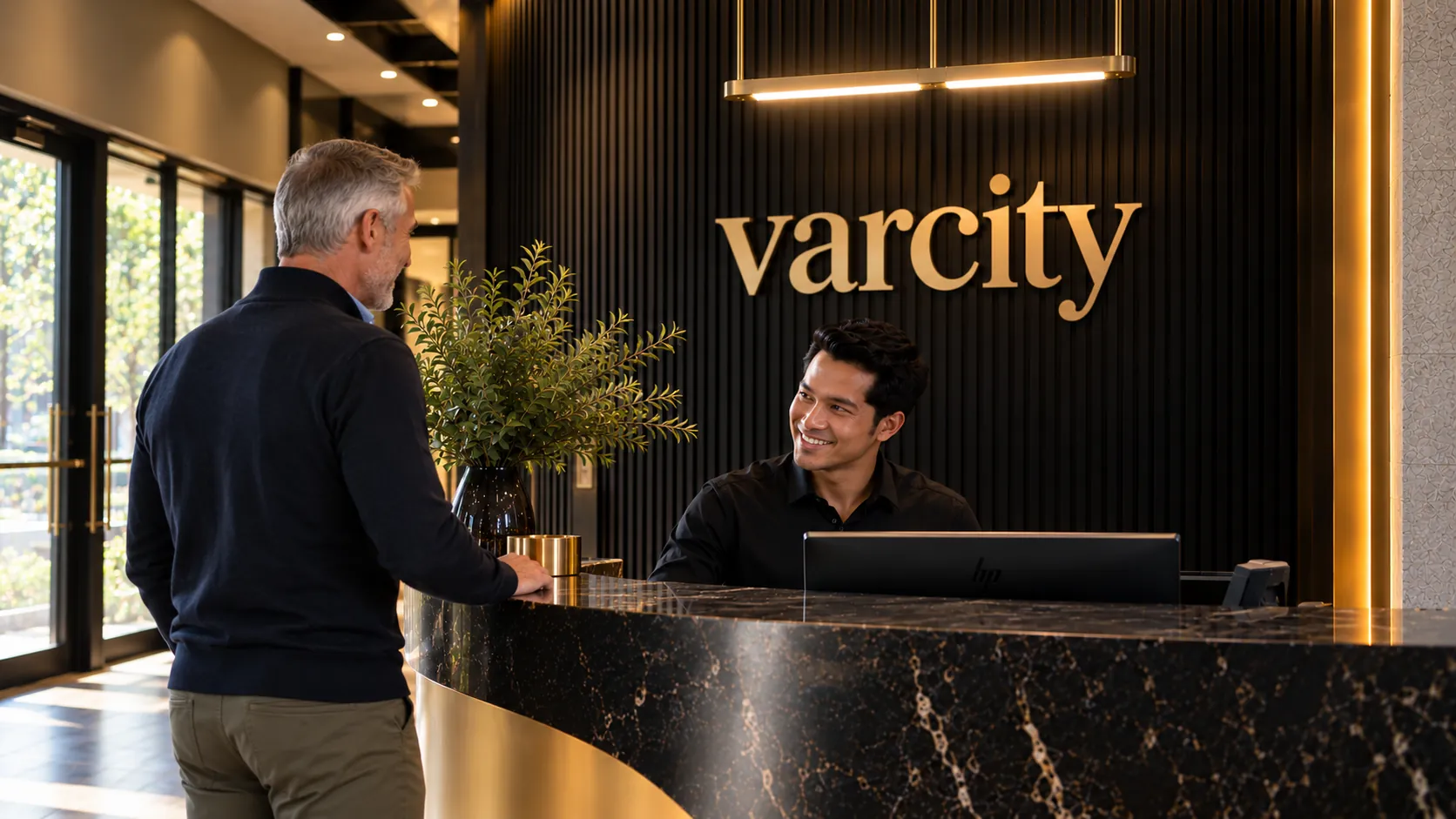 A smiling Varcity concierge chatting with a resident across the black marble and brass front desk, beneath the gold varcity sign